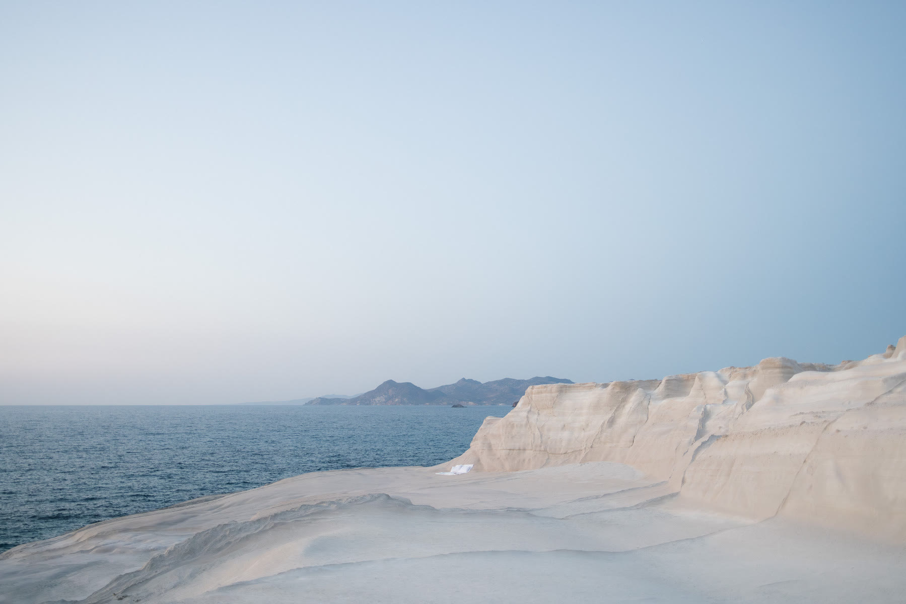 A serene coastal scene on the Greek Island of Milos features a minimalist setup with a sheet and pillow on a rocky shoreline overlooking calm waters and distant islands.